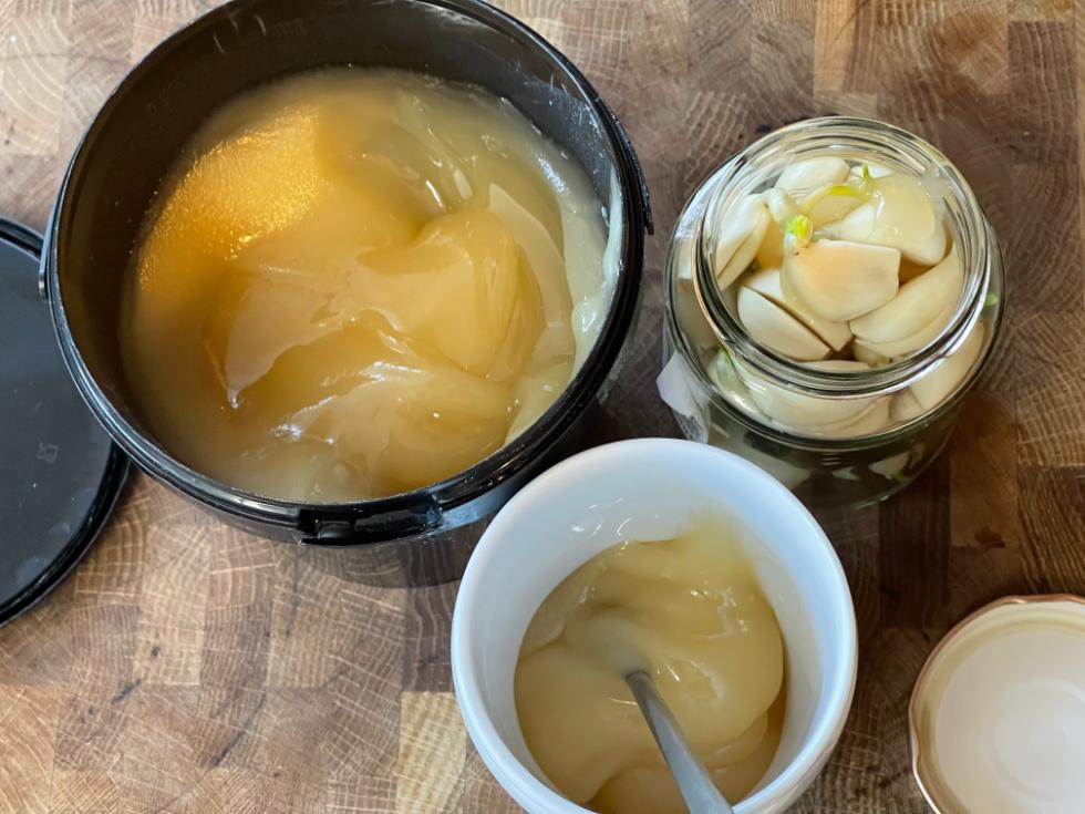 Bowls of raw honey beside a small jar of garlic fermented in honey with fresh garlic cloves on a rustic wooden board.